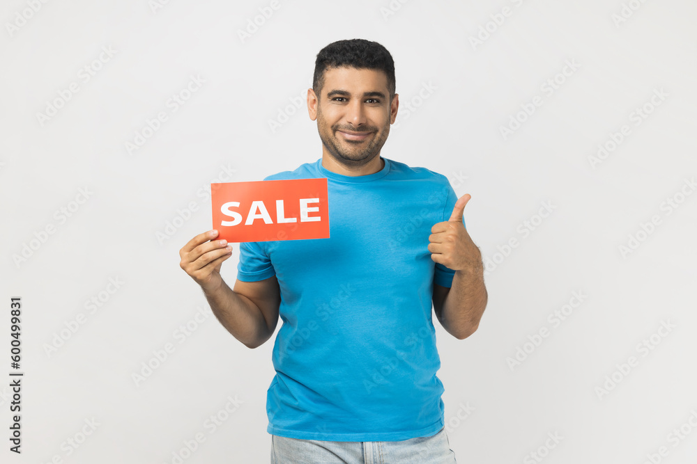 Portrait of cheerful joyful man wearing blue T- shirt standing holding sale card in hands looking at camera with toothy smile, showing thumb up. Indoor studio shot isolated on gray background. Portrait of cheerful joyful man wearing blue T- shirt standing holding sale card in hands looking at camera with toothy smile, showing thumb up. Indoor studio shot isolated on gray background.