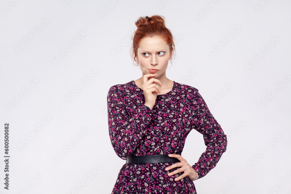 Portrait of redhead woman wearing dress holding chin and pondering idea, planning strategy, musing and solving difficult question in mind. Indoor studio shot isolated on gray background. Portrait of redhead woman wearing dress holding chin and pondering idea, planning strategy, musing and solving difficult question in mind. Indoor studio shot isolated on gray background.