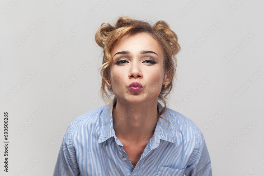 Air kiss for you. Portrait of blonde woman keeps lips folded, expressing love, blowing mwah, having romantic expression, wearing blue shirt. Indoor studio shot isolated on gray background. Air kiss for you. Portrait of blonde woman keeps lips folded, expressing love, blowing mwah, having romantic expression, wearing blue shirt. Indoor studio shot isolated on gray background.