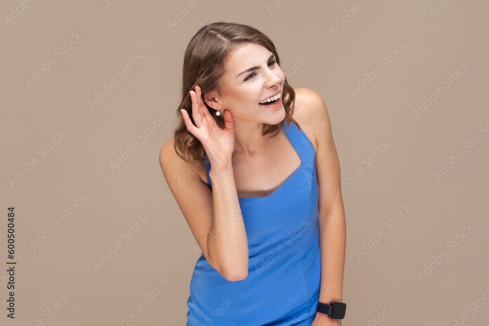 Portrait of attractive beautiful young adult woman with wavy hair standing palm near ear, listening private conversation, wearing blue dress. Indoor studio shot isolated on light brown background. Portrait of attractive beautiful young adult woman with wavy hair standing palm near ear, listening private conversation, wearing blue dress. Indoor studio shot isolated on light brown background.