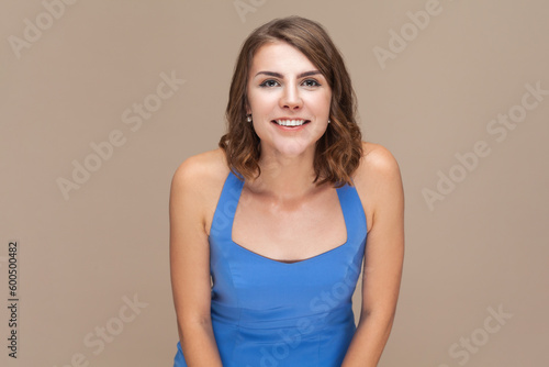 Portrait of smiling positive happy woman with wavy hair looking at camera with toothy smile, being in good mood, being glad, wearing blue dress. Indoor studio shot isolated on light brown background.