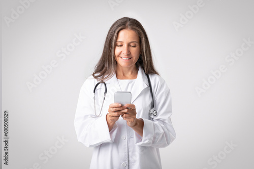 Glad senior woman therapist in white coat with stethoscope using smartphone for work  light studio background