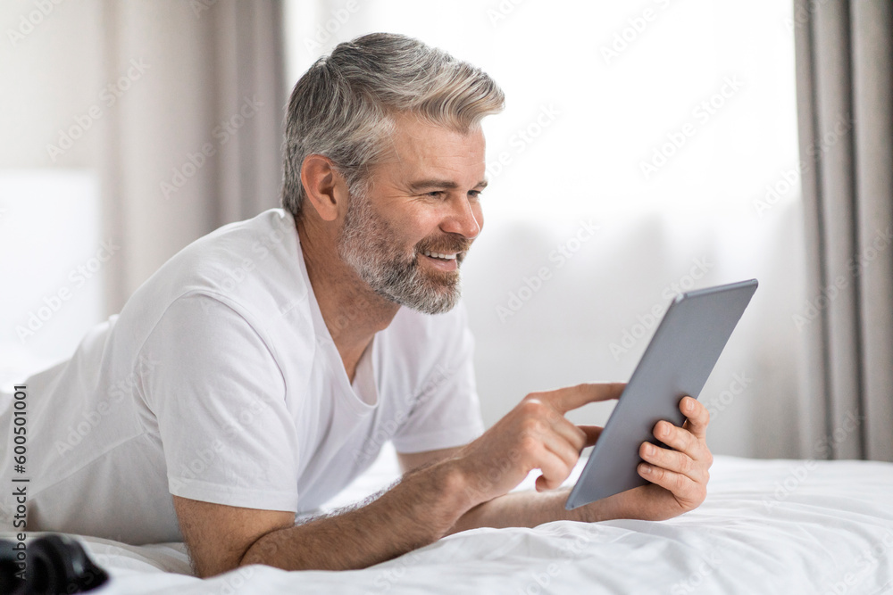 Joyful middle aged man chilling in bed, using digital tablet