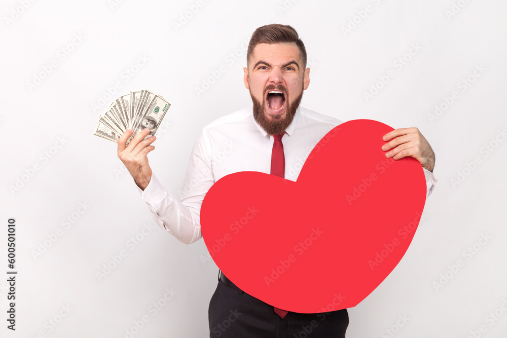 Portrait of angry bearded man wearing white shirt and tie, showing big red heart and dollar banknotes, screaming with hate and aggression. Indoor shot isolated on gray background.