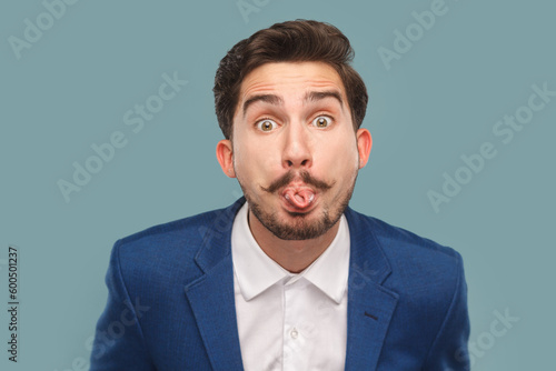 Portrait of funny handsome man with mustache standing sticking tongue out, showing childish behavior, wearing white shirt and jacket. Indoor studio shot isolated on light blue background.