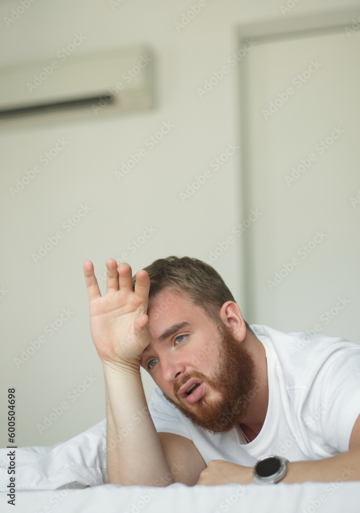 Young bearded man using air conditioner at home, lay on bed cooling off during hot weather, suffering from heat, high temperature. Broken AC. Vertical photo. Young bearded man using air conditioner at home, lay on bed cooling off during hot weather, suffering from heat, high temperature. Broken AC. Vertical photo.