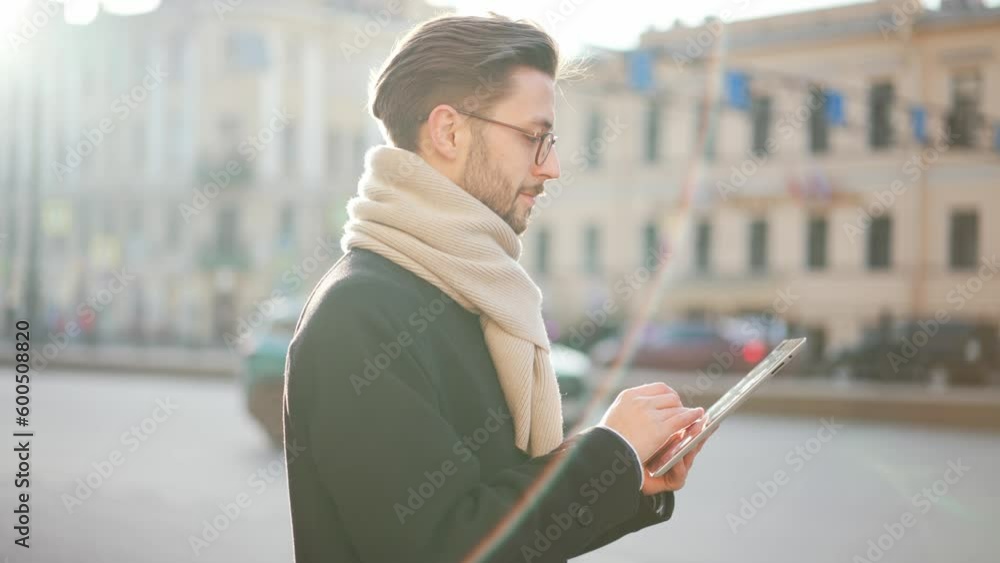 Young handsome man in glasses with beard stands on busy street at back moving cars in city center and uses digital tablet. Successful businessman online works on freelance. Caucasian face portrait. Young handsome man in glasses with beard stands on busy street at back moving cars in city center and uses digital tablet. Successful businessman online works on freelance. Caucasian face portrait.