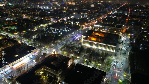A big concert in the night city. Drone view of a crowd of people, rays and lights of light. A large building with lighting. There are roads with cars, pedestrian streets with people and a park around