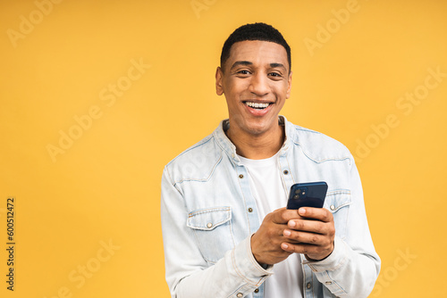 Smiling african american young man holding phone isolated on yellow studio background with copy space aside, african guy using mobile applications, texting.