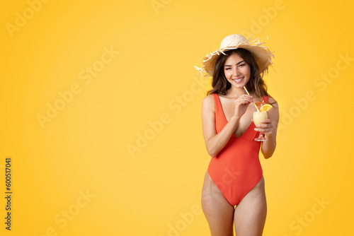 Smiling millennial asian woman in swimsuit and hat with tropical cocktail, enjoy free time #600517041