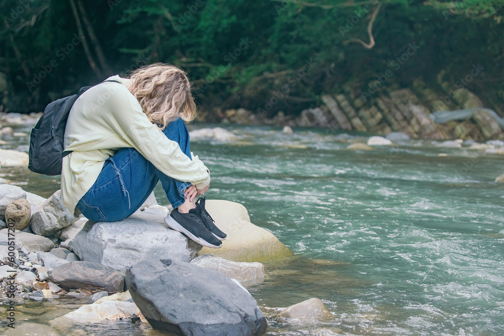 frustrated, tired tourist girl sits alone on the bank of a mountain river on the rocks, she clasped her legs in her hands and laid her head on them during a hike in nature frustrated, tired tourist girl sits alone on the bank of a mountain river on the rocks, she clasped her legs in her hands and laid her head on them during a hike in nature