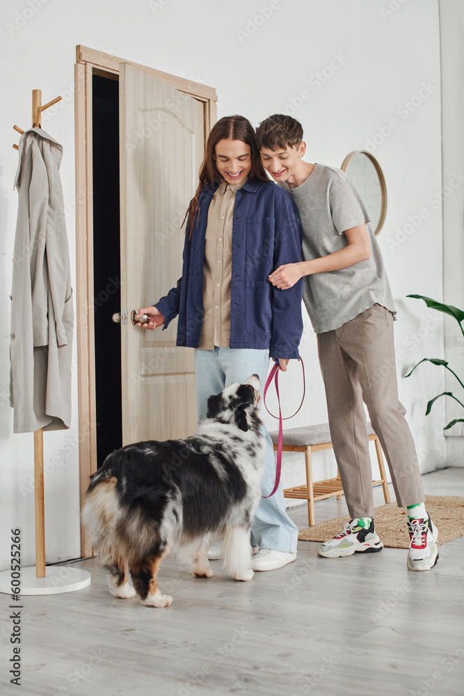 happy gay couple in casual outfits standing in modern hallway next to coat rack and looking at Australian shepherd dog with leash and smiling together at home happy gay couple in casual outfits standing in modern hallway next to coat rack and looking at Australian shepherd dog with leash and smiling together at home