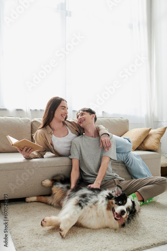 cheerful gay man sitting on carpet and cuddling Australian shepherd dog and looking at happy partner with long hair holding book in modern living room © LIGHTFIELD STUDIOS