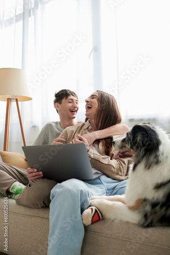 cheerful gay partners in casual clothes laughing while hugging each other and sitting together near laptop and cute furry friend resting near them on couch in modern living room