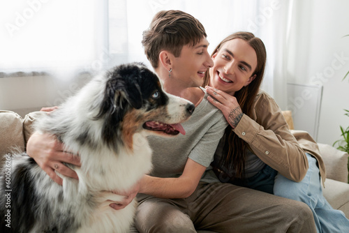 Cheerful and long haired gay man with tattoo on hand hugging and looking at young boyfriend near blurred Australian shepherd dog sitting on couch in living room at home
