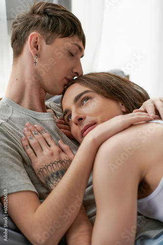 Young homosexual man in sleepwear kissing tattooed and long haired boyfriend looking away while resting in bedroom at home in morning time