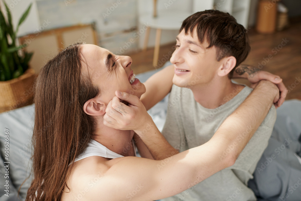 Blurred homosexual man in sleepwear touching cheek of cheerful long haired boyfriend in pajama resting on blurred bed in modern house in morning Blurred homosexual man in sleepwear touching cheek of cheerful long haired boyfriend in pajama resting on blurred bed in modern house in morning