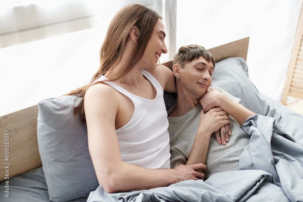 Overjoyed and long haired homosexual man in pajama hugging pleased young boyfriend with closed eyes while sitting on bed in morning time at home Overjoyed and long haired homosexual man in pajama hugging pleased young boyfriend with closed eyes while sitting on bed in morning time at home