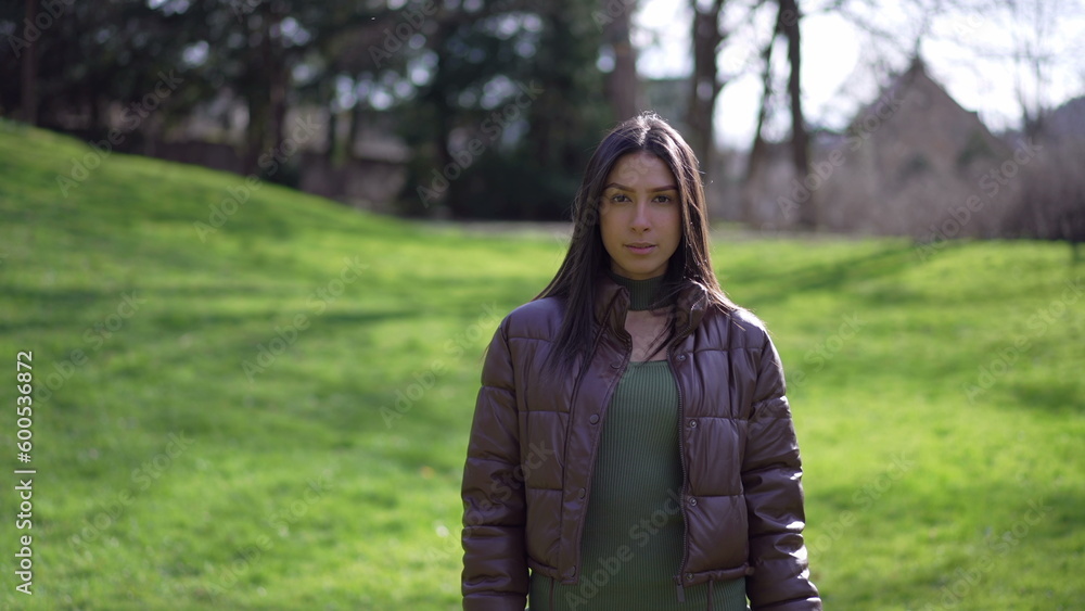 One young Middle Eastern woman standing outdoors at park looking at camera with neutral expression One young Middle Eastern woman standing outdoors at park looking at camera with neutral expression