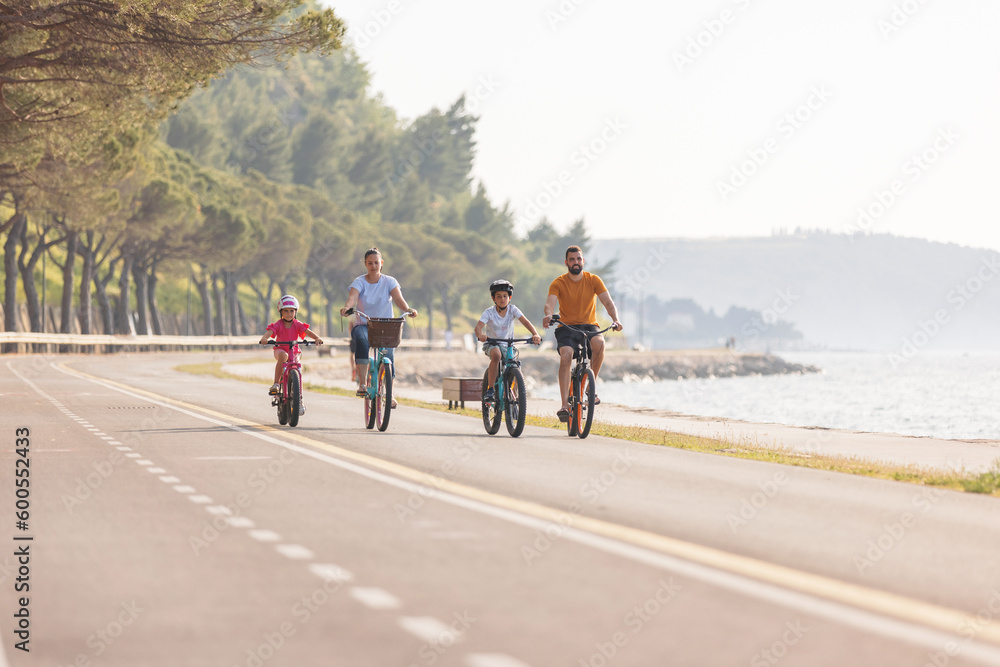 Obraz premium Two kids, with helmets on their heads, and smiling parents riding bikes on a family-friendly cycle route along a sea coastline, front view.
