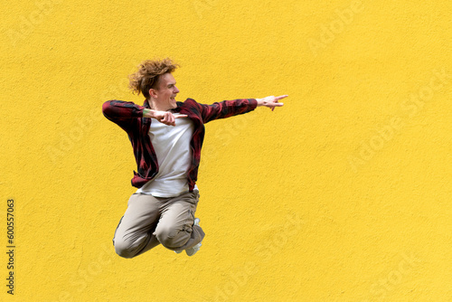 young guy student jumps against the background of yellow isolated wall and shows his hands to the side on copy space