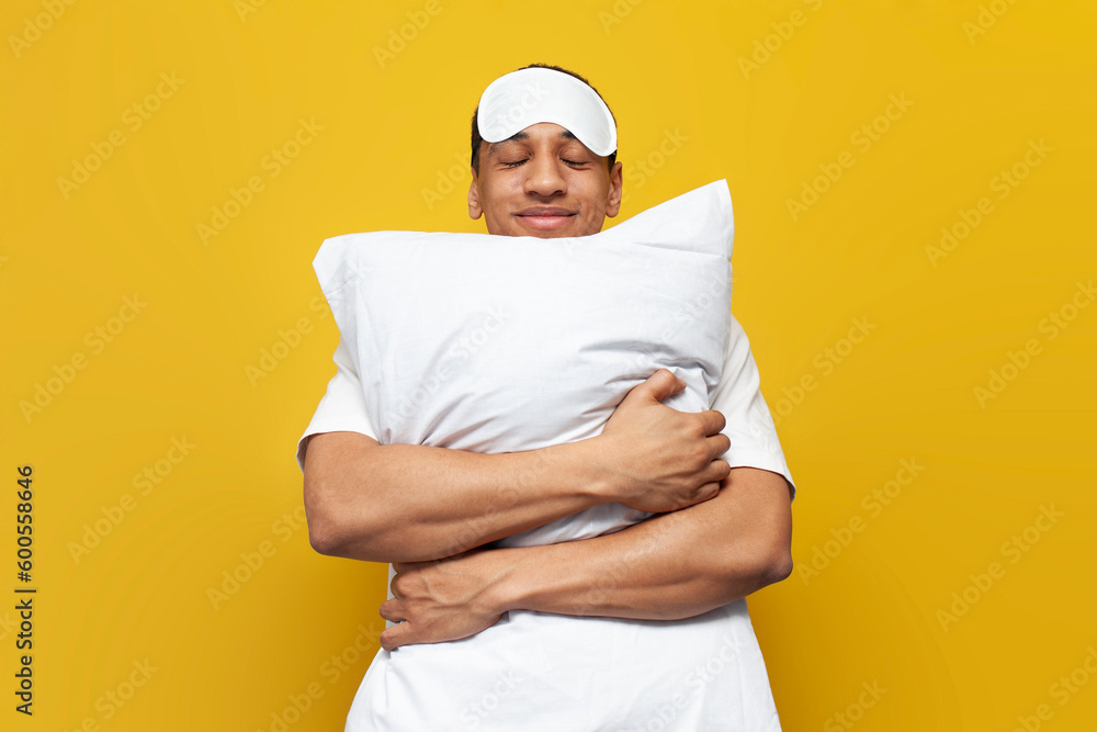 young african american guy in pajamas and sleep mask hugs soft comfortable pillow and smiles on yellow background young african american guy in pajamas and sleep mask hugs soft comfortable pillow and smiles on yellow background