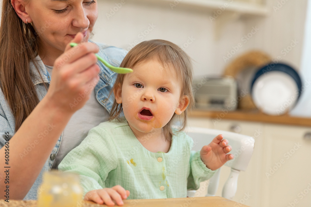 Happy family at home. Mother feeding her baby girl from spoon in kitchen. Little toddler child with messy funny face eats healthy food at home. Young woman mom giving food to kid daughter Happy family at home. Mother feeding her baby girl from spoon in kitchen. Little toddler child with messy funny face eats healthy food at home. Young woman mom giving food to kid daughter