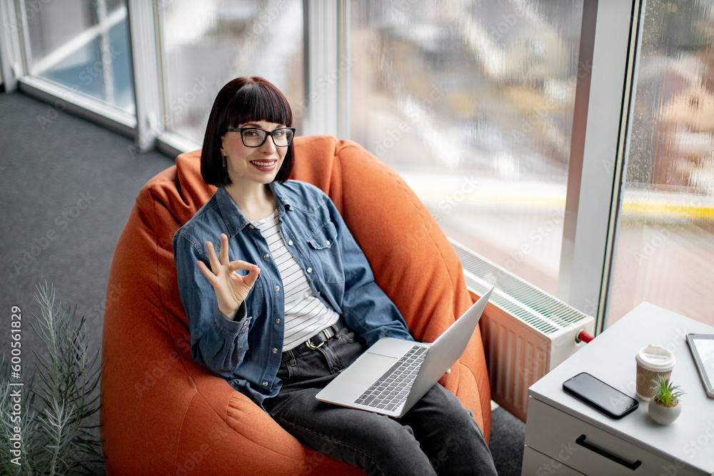 Caucasian lady with short hair wearing denim jacket and black jeans sitting on chair and smiling at camera. Attractive female using wireless laptop for distant work at modern coworking space. Caucasian lady with short hair wearing denim jacket and black jeans sitting on chair and smiling at camera. Attractive female using wireless laptop for distant work at modern coworking space.