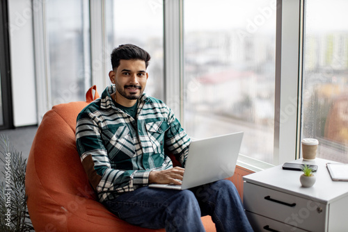 Portrait of nice-looking indian employee sitting on soft office chair with personal laptop on knees and smiling at camera. Imposing dark haired man taking short break after intensive working day.