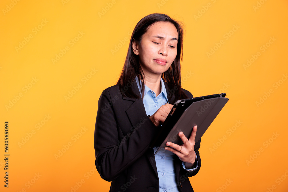 Asian woman browsing information on tablet computer, working at research project in studio over yellow background. Cheerful model using electronic gadget with touchscreen for creative work. Asian woman browsing information on tablet computer, working at research project in studio over yellow background. Cheerful model using electronic gadget with touchscreen for creative work.