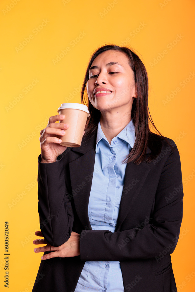 Satisfied asian model holding cup of coffee drinking hot beverage enjoying free time before start to photoshot in studio. cheerful young adult with brunette long hair standing over yellow background Satisfied asian model holding cup of coffee drinking hot beverage enjoying free time before start to photoshot in studio. cheerful young adult with brunette long hair standing over yellow background
