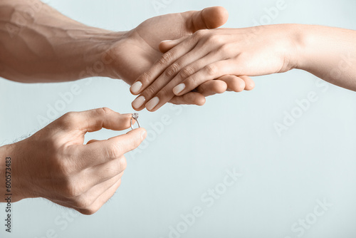 Man putting engagement ring on woman's finger against light background, closeup