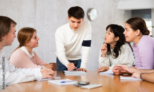 Cheerful young student discussing learning assignment with friendly coursemates gathered in classroom