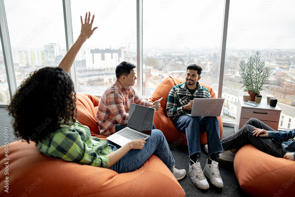 Smiling indian male holding electronic device on knees while active female raising hand for question in modern meeting room. Multiethnic business colleagues being engaged in brief conference indoors. Smiling indian male holding electronic device on knees while active female raising hand for question in modern meeting room. Multiethnic business colleagues being engaged in brief conference indoors.