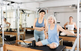 Focused positive elderly woman practicing pilates on reformer to improve and maintain mobility under supervision of attentive young Asian female trainer..