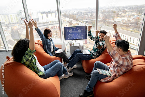 Excited multiracial businesspeople raising hands cheerfully while sitting with laptops and tablet on terracotta poufs in creative office. Automotive engineers celebrating design project completion.
