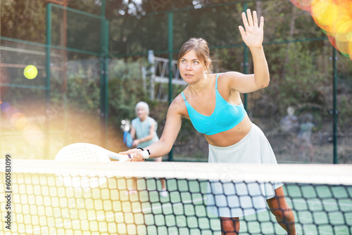 Happy woman learning to play padel game on tennis court outdoor. Other athletes are training in the background © JackF