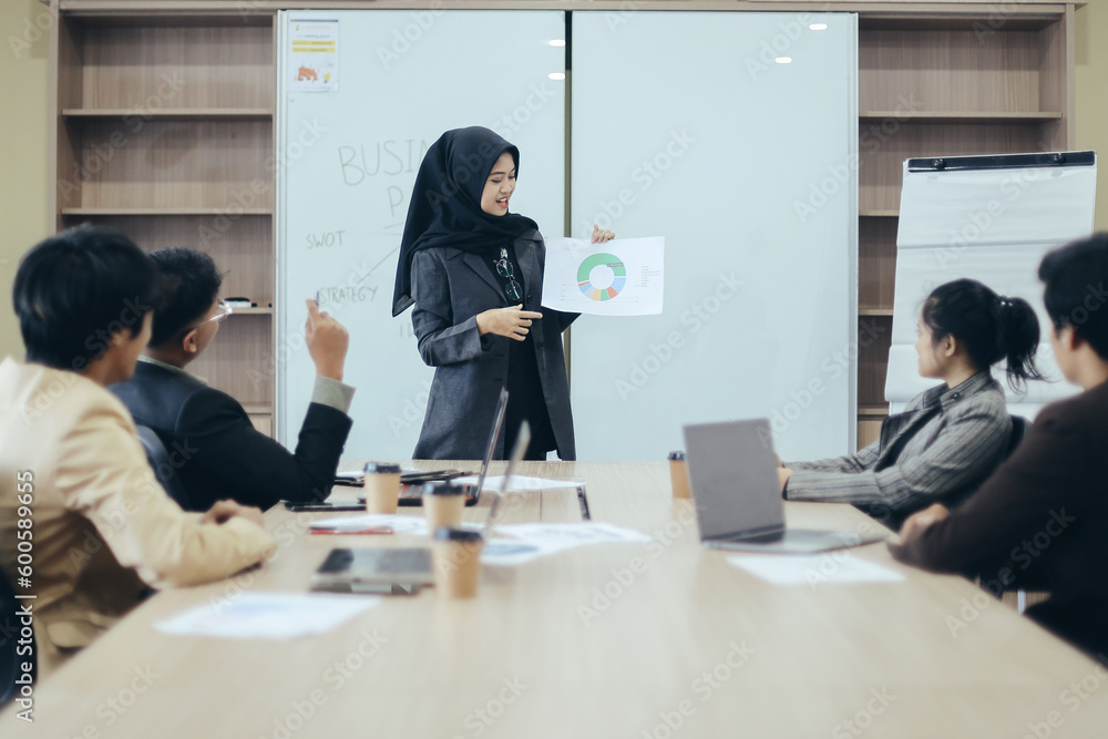 Fototapeta premium Asian muslim woman having presentation to team showing sales graphic at the office. Businesswoman lead group of young Asian business creative team in brainstorm meeting presentation.
