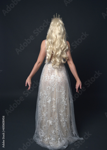 Full length portrait of beautiful women with long blonde hair, wearing fantasy  princess crown and elegant white ball gown, standing pose with hand gesture. Isolated on dark grey studio background.