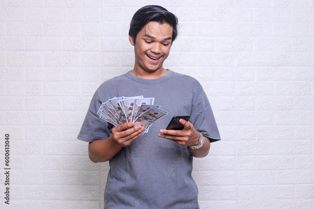 Young Asian man holding money showing excited expression when looking to his phone