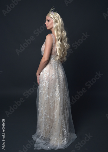 Full length portrait of beautiful women with long blonde hair, wearing fantasy princess crown and elegant white ball gown, standing pose with hand gesture. Isolated on dark grey studio background.