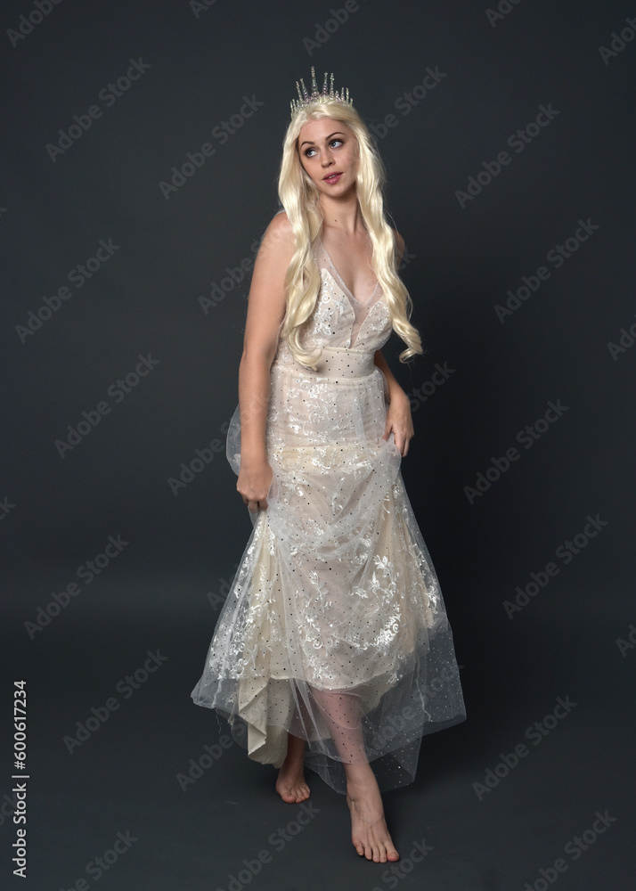 Full length portrait of beautiful women with long blonde hair, wearing fantasy princess crown and elegant white ball gown, standing pose with hand gesture. Isolated on dark grey studio background. Full length portrait of beautiful women with long blonde hair, wearing fantasy princess crown and elegant white ball gown, standing pose with hand gesture. Isolated on dark grey studio background.