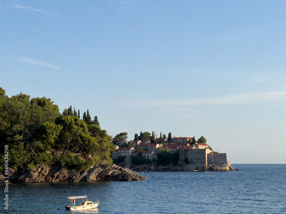 Small boat sails to the island of Sveti Stefan, skirting a rocky hill covered with green trees. Montenegro