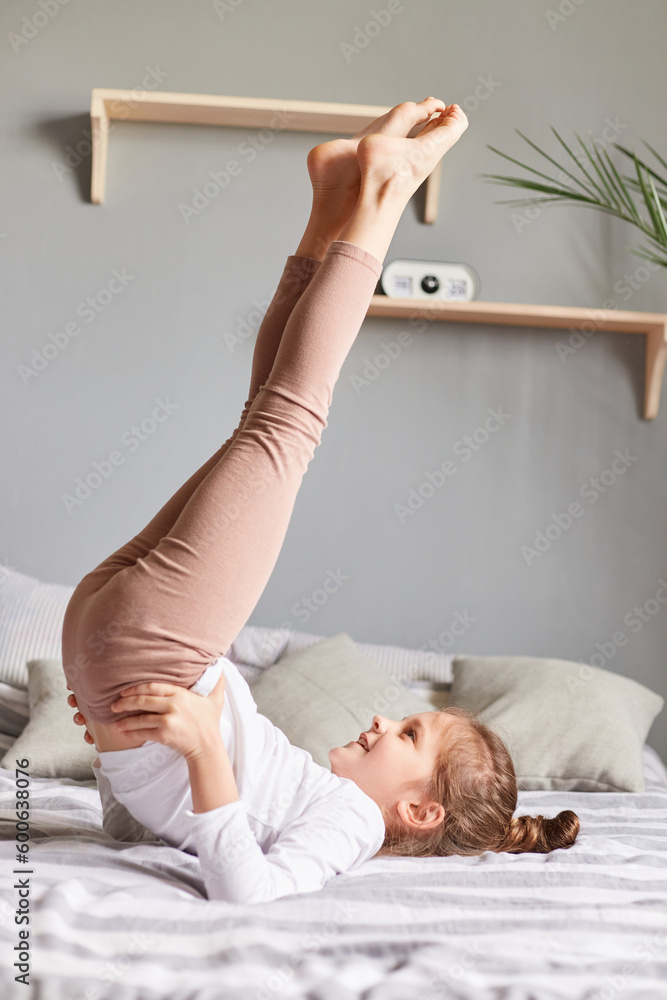 Cute little girl doing workout in bedroom at home on bad having fun raised her legs keeps hands in hips doing exercise birch in bedroom morning training and stretching. Stock Photo