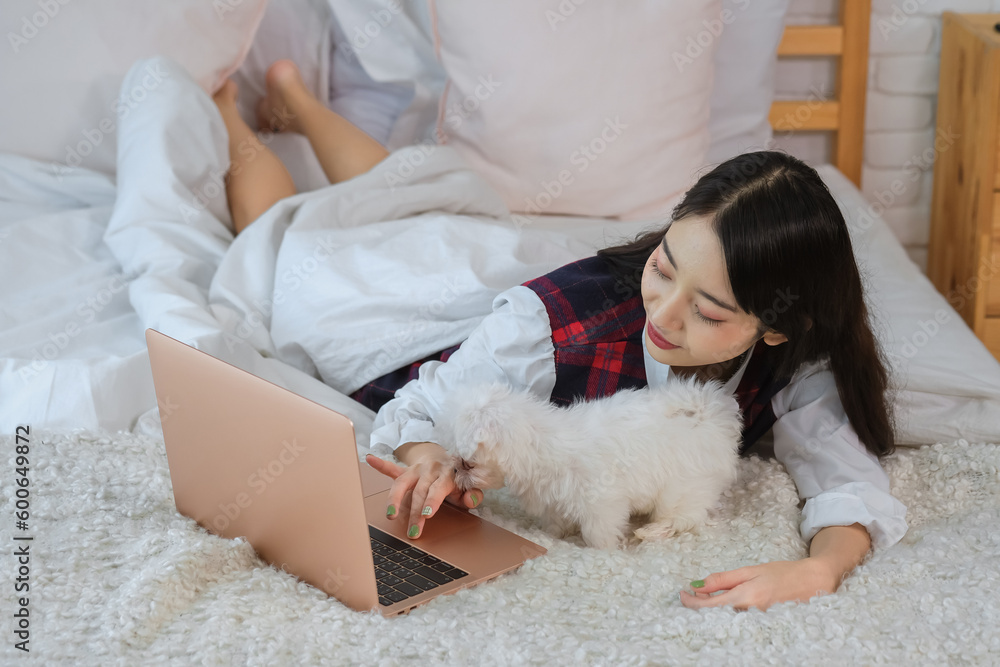 Beautiful asian woman seemed relaxed and content as she worked on her laptop while holding her cute white dog on the bed in her cozy bedroom. Beautiful asian woman seemed relaxed and content as she worked on her laptop while holding her cute white dog on the bed in her cozy bedroom.