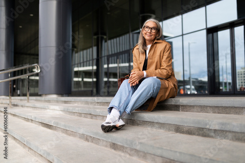 confident mature adult female accountant with gray hair sitting on steps of business center