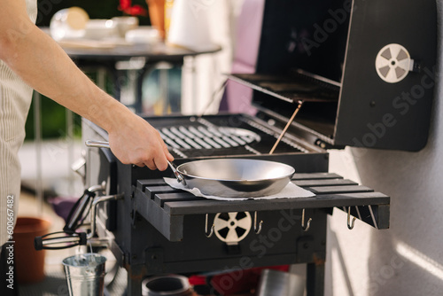 Close-up of man in apron take frying pan from side stand on BBQ grill on terrace 
