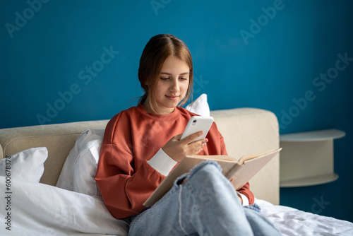 Teen girl reading book photographing page on smartphone to share in social media sitting in bed at home. Caucasian teenager bookworm nerd kinds of literature. Leisure, taking picture on mobile phone. photo