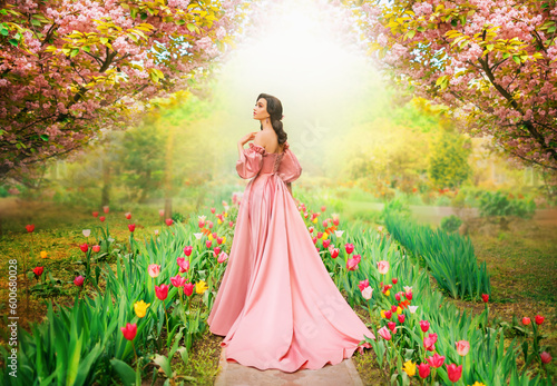 Fantasy girl princess walks in blooming spring garden flowers tulips sakura tree green grass old alley. Woman queen in long luxurious royal pink dress with train puffed sleeves vintage style art photo