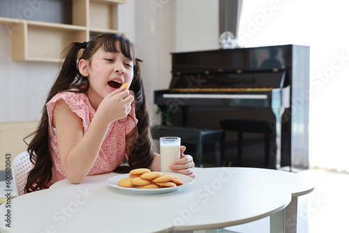 Kid asian black hair girl sitting on table and drinking milk while eating vanilla cookies for breakfast with enjoying time. Tasty food and delicious food with happy meal lifestyle kid concept